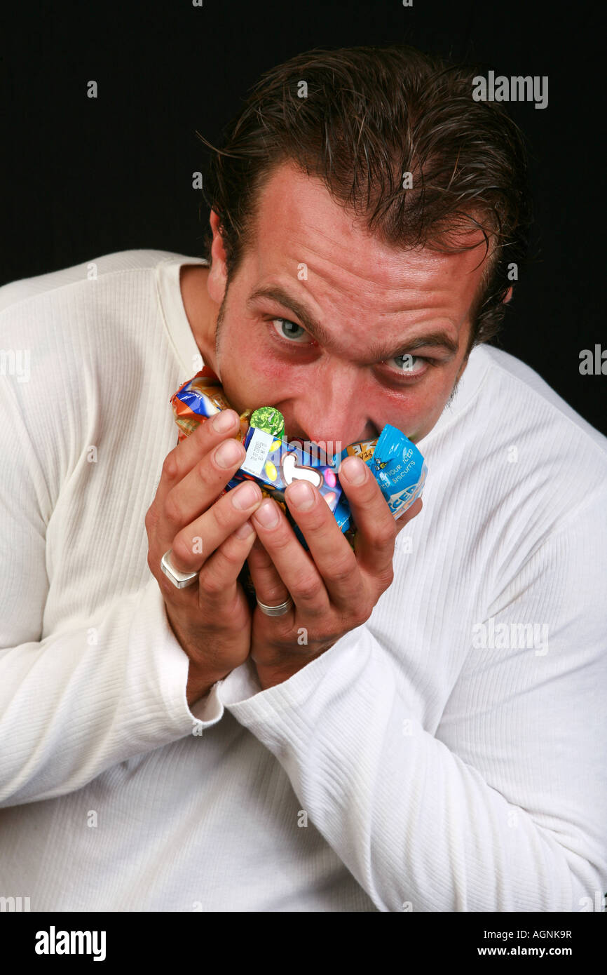 Jeune homme 20s bonbons bonbons chocolats farce grignotines sucrées dans sa bouche avide de la malbouffe addiction mauvaise alimentation Banque D'Images