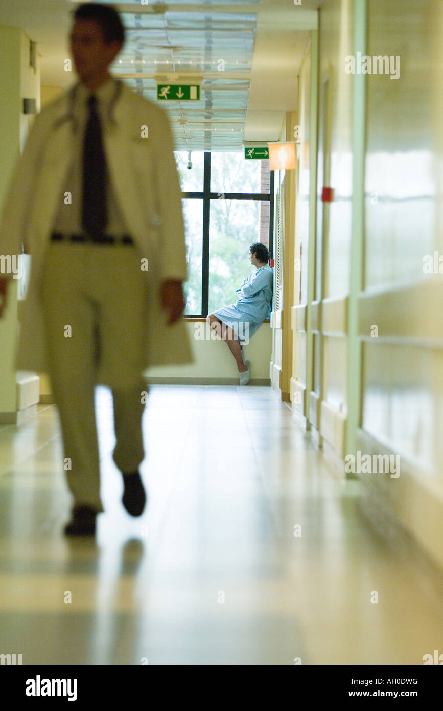 Homme assis, looking out window, portant chemise d'hôpital, médecin en premier plan de marche Banque D'Images