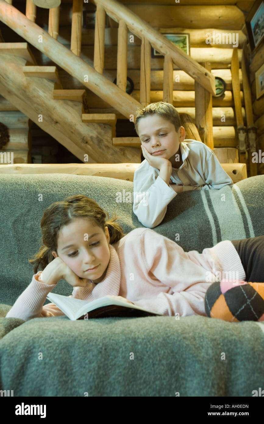 Girl on couch reading book, boy looking over shoulder Banque D'Images