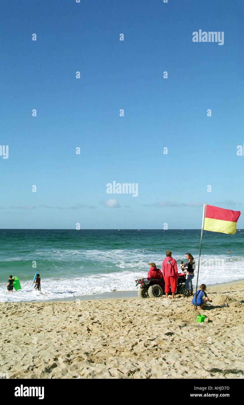Scène de plage avec des bodyboarders, un enfant construisant un château de sable, un maître-nageur assis sur un quad et un autre maître-nageur parlant à une fille. Banque D'Images