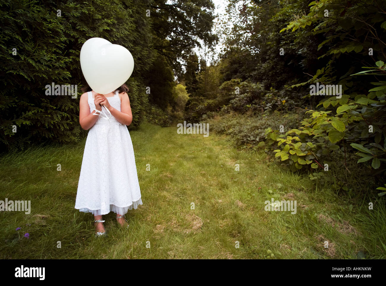 Enfant Jeune fille en robe blanche holding white heart shaped balloon sur son visage dans le jardin envahi par la Banque D'Images