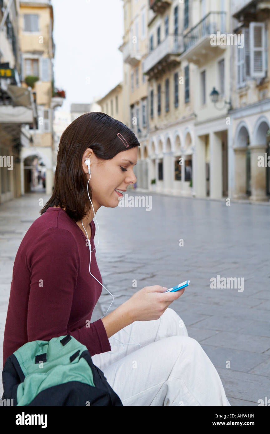 Jeune fille assise sur les marches de la rue dans les oreilles des écouteurs de MP3 dans la main smiling vieille ville en arrière-plan. Banque D'Images