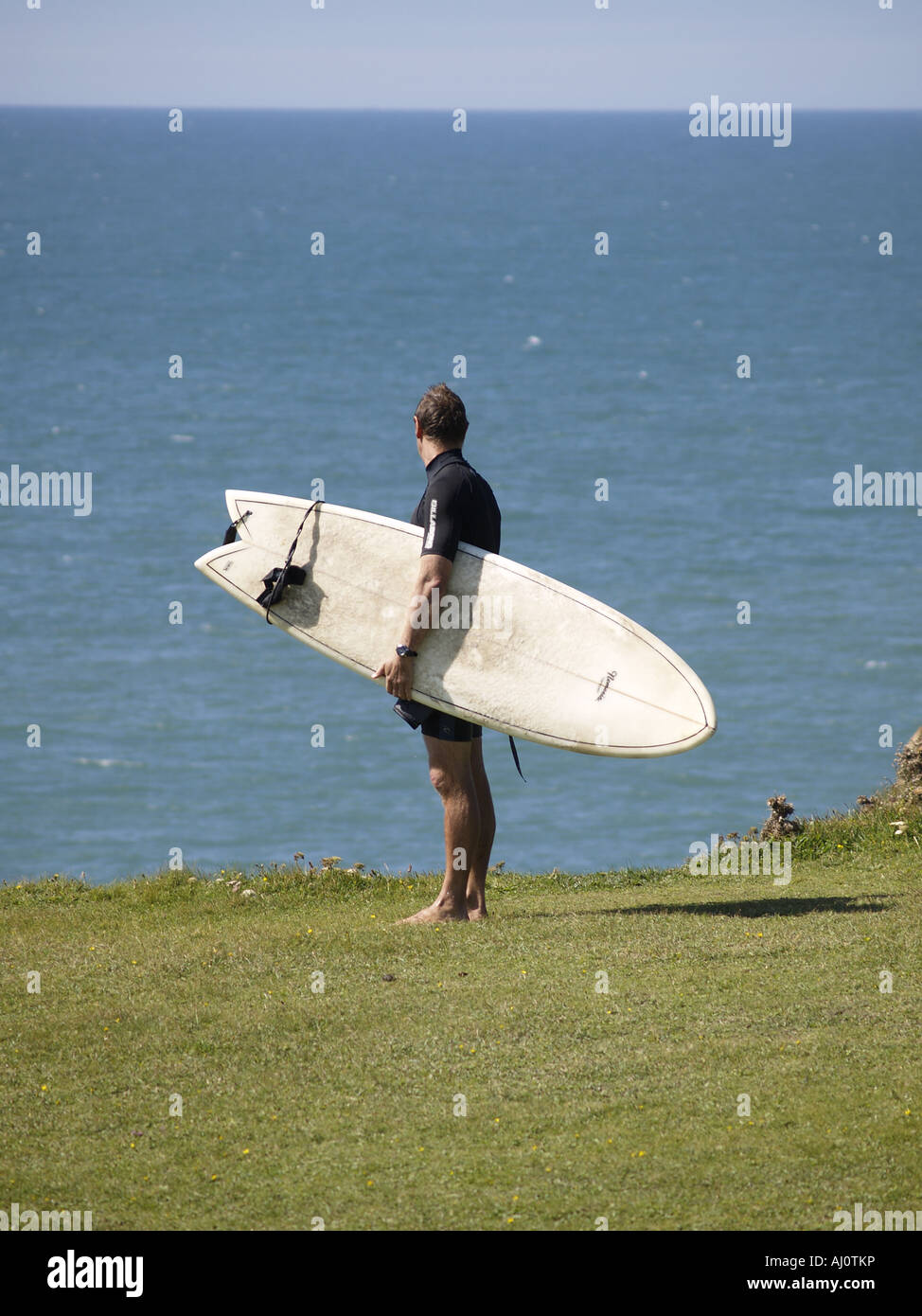 Surfer debout sur la falaise au-dessus de la plage de Crooklets à Bude, une planche de surf sous le bras, en regardant les vagues. Banque D'Images