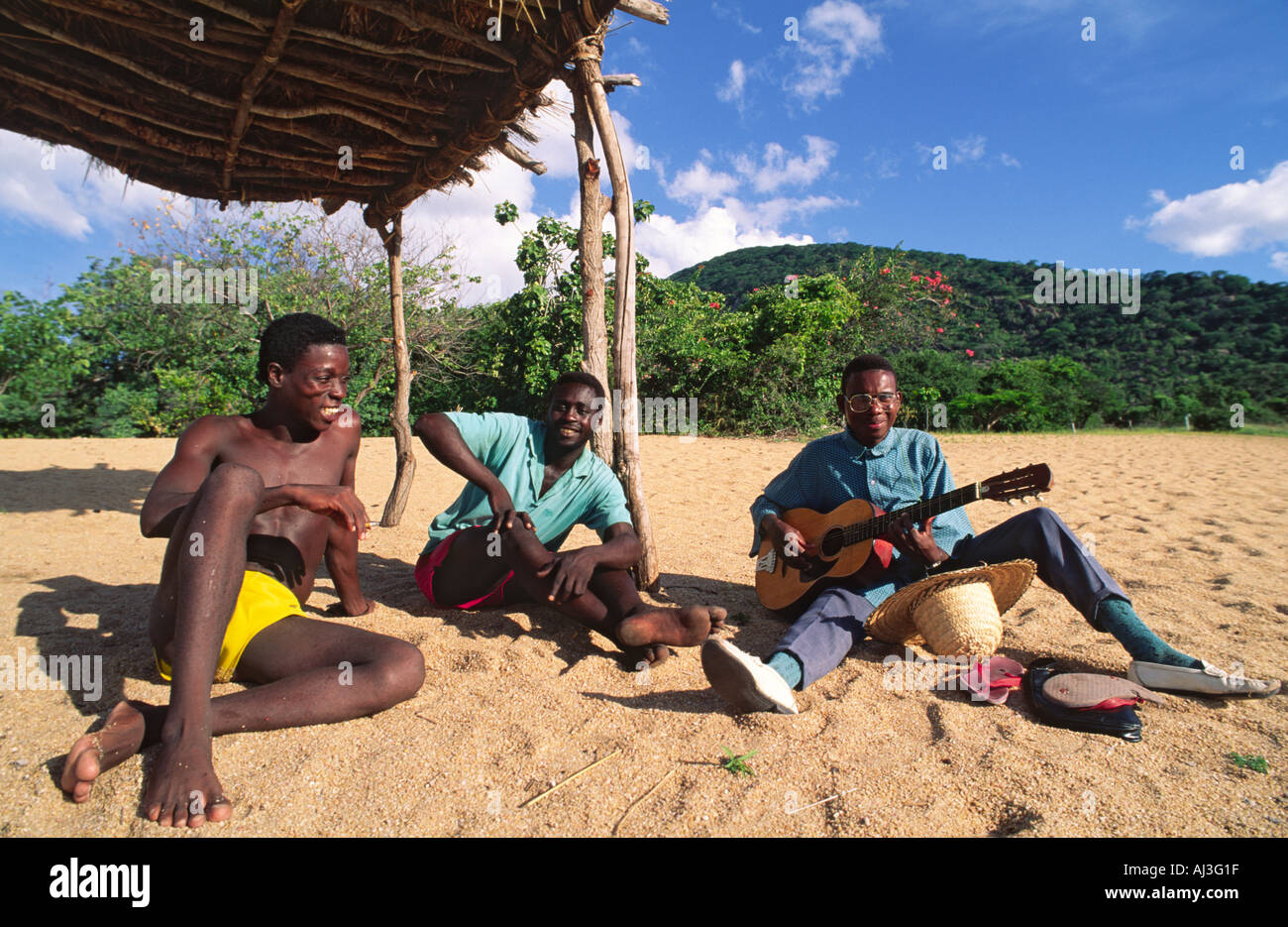 Deux gars du Malawi et de détente de l'écoute d'un jeune ami jouer de la guitare sur une plage sur le lac Malawi, Malawi. Banque D'Images