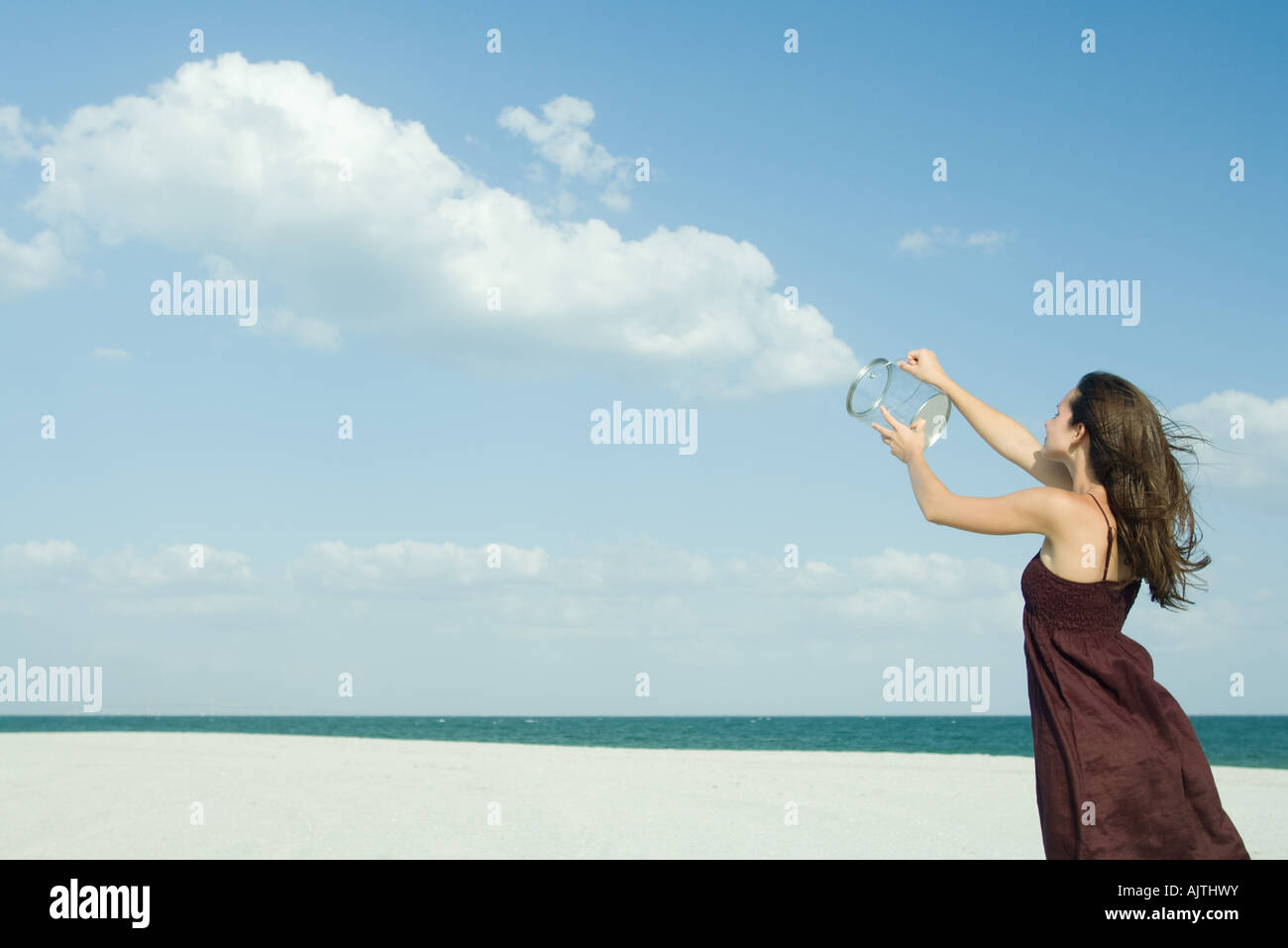 Woman standing on beach, holding up contenant transparent, libérant des nuages, illusion optique Banque D'Images