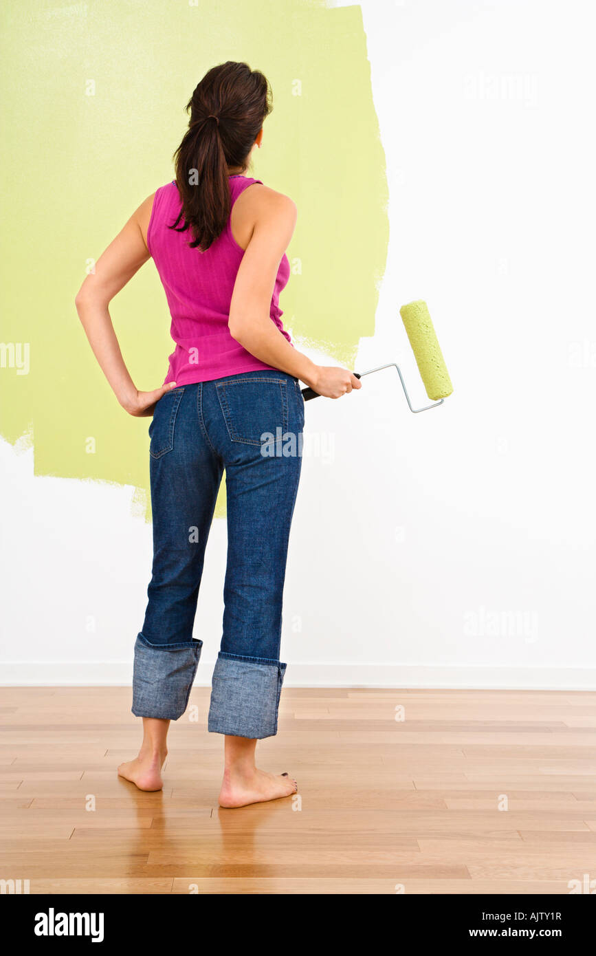 Woman holding paint roller admirant partiellement peint mur intérieur Banque D'Images