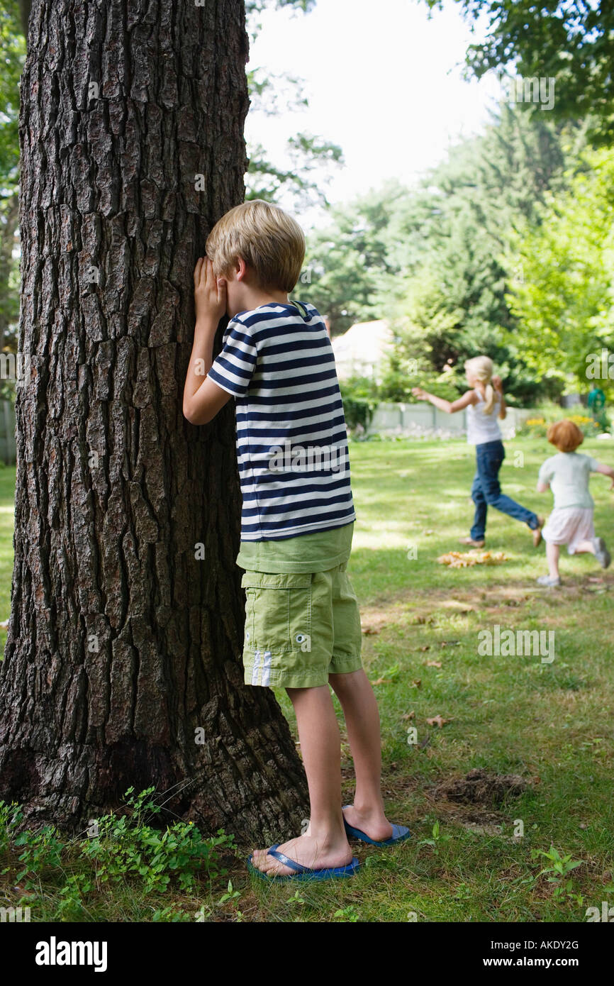 Boy (5-6) Comité permanent par arbre avec les yeux couverts, les enfants en cours d'exécution en arrière-plan Banque D'Images