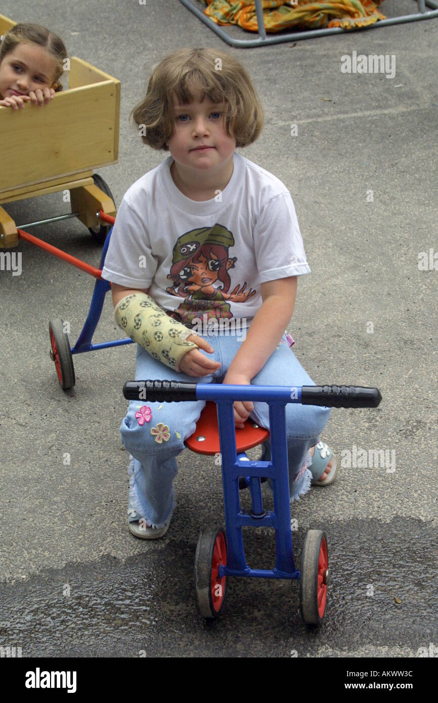 Fille de pépinière sur tricylce dans les jardins d'aire de jeux avec le bras dans un plâtre Banque D'Images
