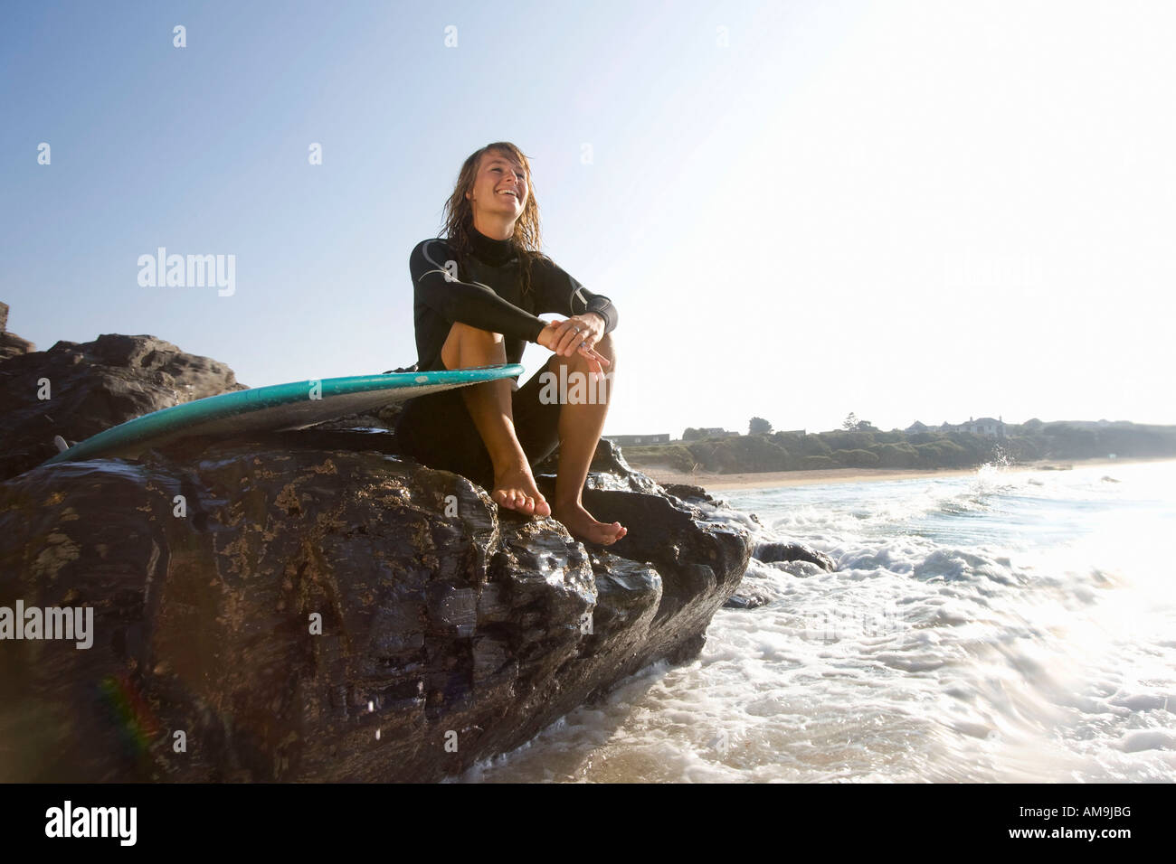 Femme assise sur de gros rochers avec surfboard smiling. Banque D'Images