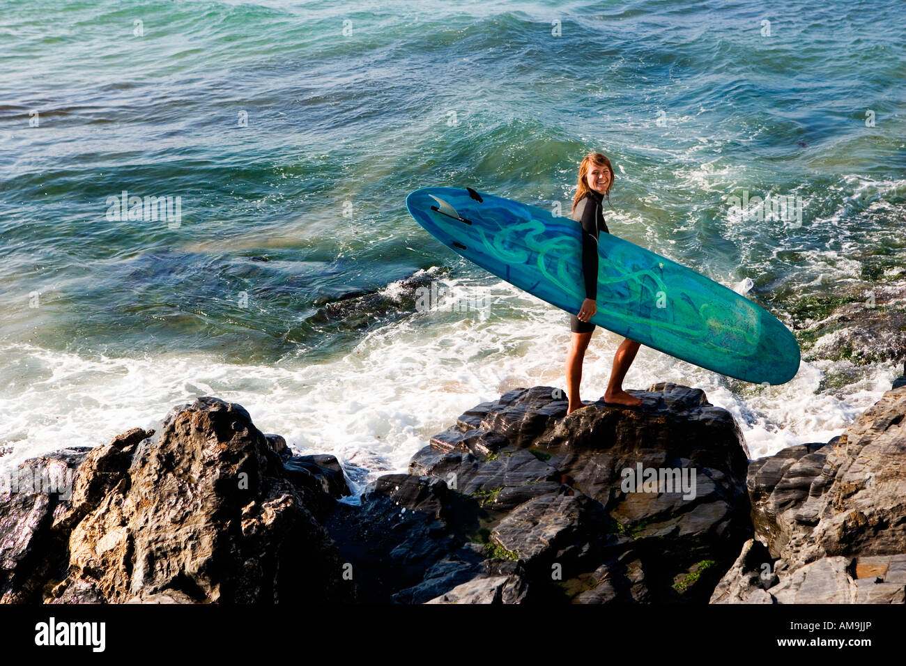 Femme debout sur de gros rochers avec un surfboard smiling. Banque D'Images