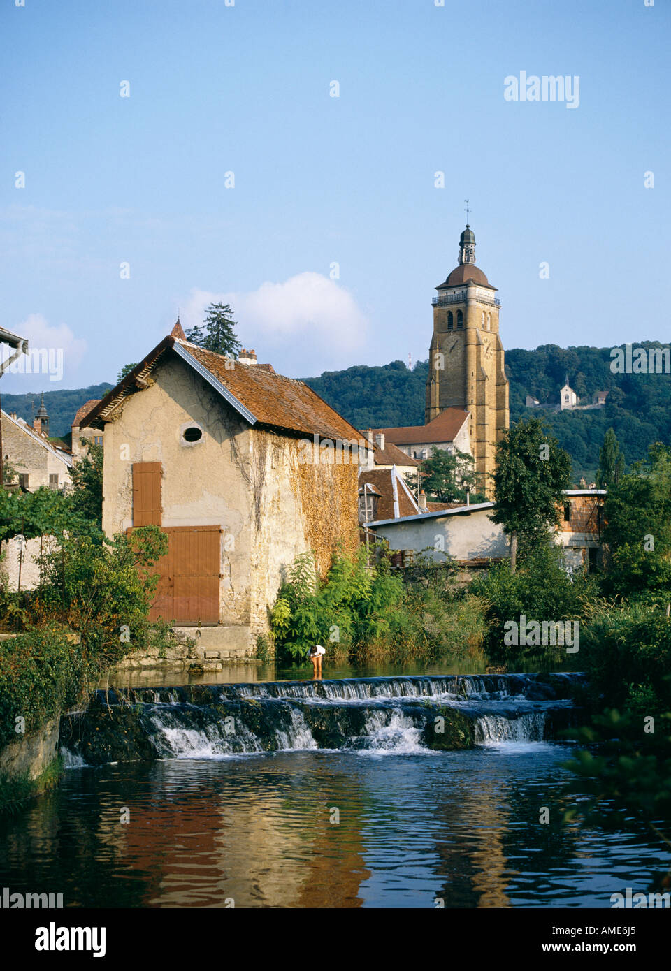 Un grand bâtiment situé au milieu des arbres sur les rives de la rivière qui coule à travers Arbois dans le Jura français avec la tour de la 12e 13e c'église de St Just s'élevant derrière Banque D'Images