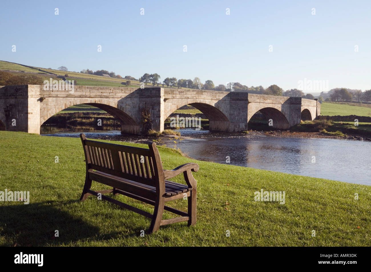 Banc vide donnant sur la rivière Wharfe et pont voûté en pierre dans village de Yorkshire Dales National Park. Burnsall Wharfedale Yorkshire Angleterre UK Banque D'Images