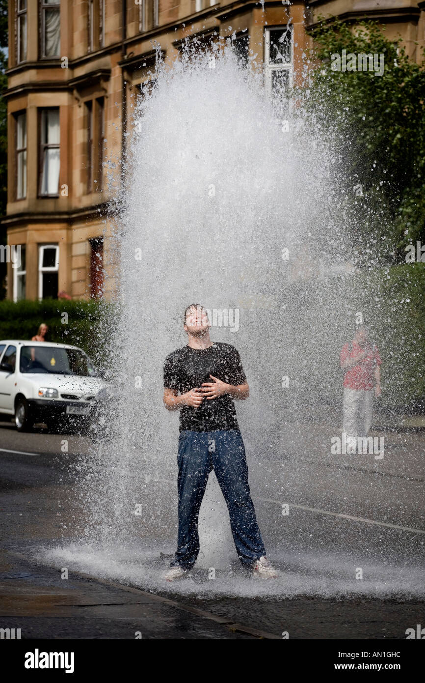 Un jeune homme se refroidit dans la chaleur de l'été comme un poteau de l'eau principale burst et jaillit de l'eau dans une rue de Glasgow Dennistoun UK Banque D'Images