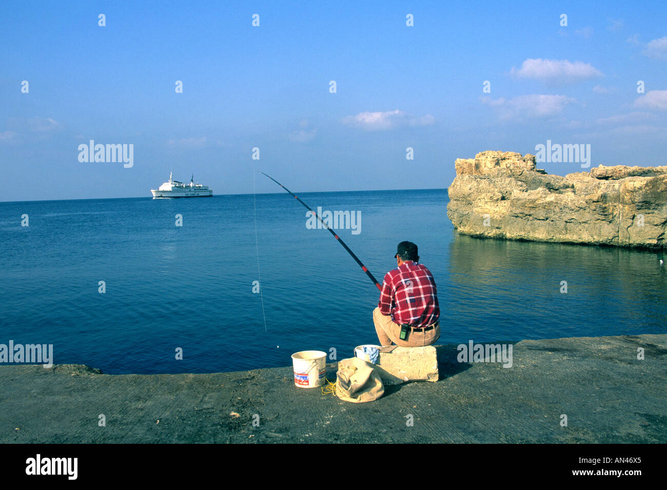 L'homme pêche dans la mer sur l'île de Gozo, Malte - avec un bateau de croisière dans l'arrière-plan Banque D'Images