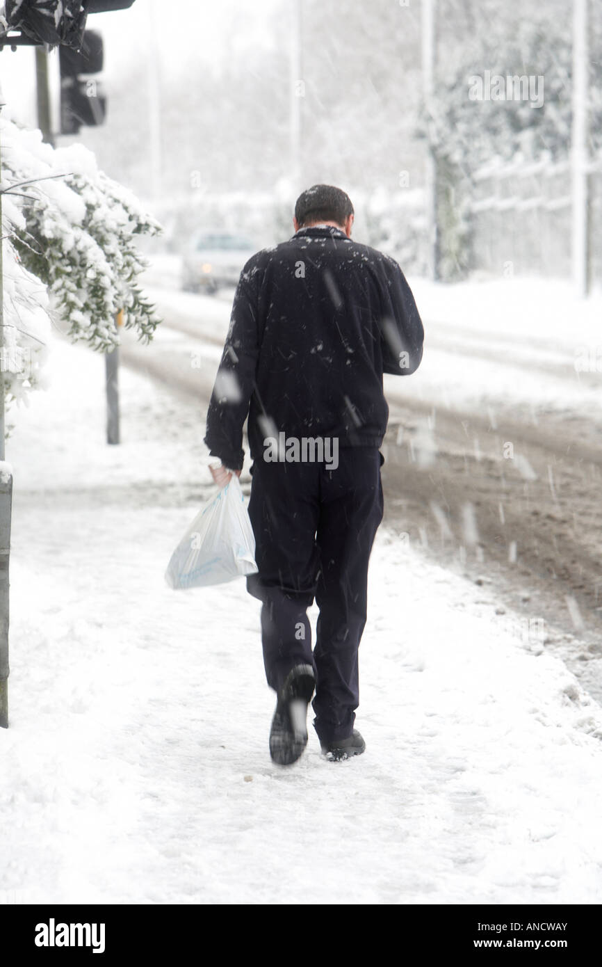 Homme sans hood marche en montée le long sentier couvert de neige transportant un sac en plastique à la recherche vers le bas au téléphone Banque D'Images