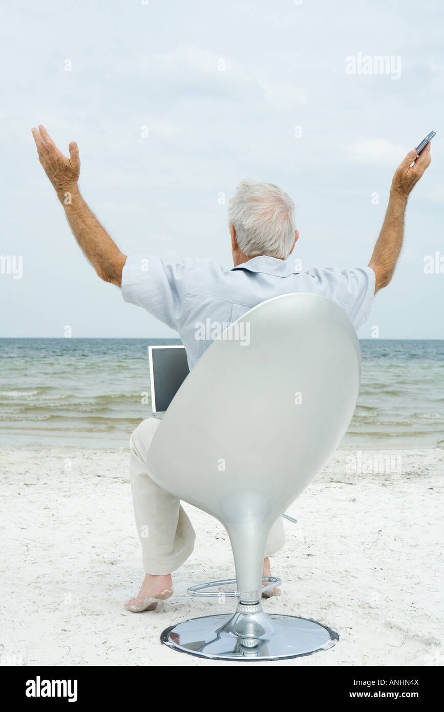 Senior man sitting in chair on beach, à l'aide d'ordinateur portable, les bras en l'air, vue arrière Banque D'Images
