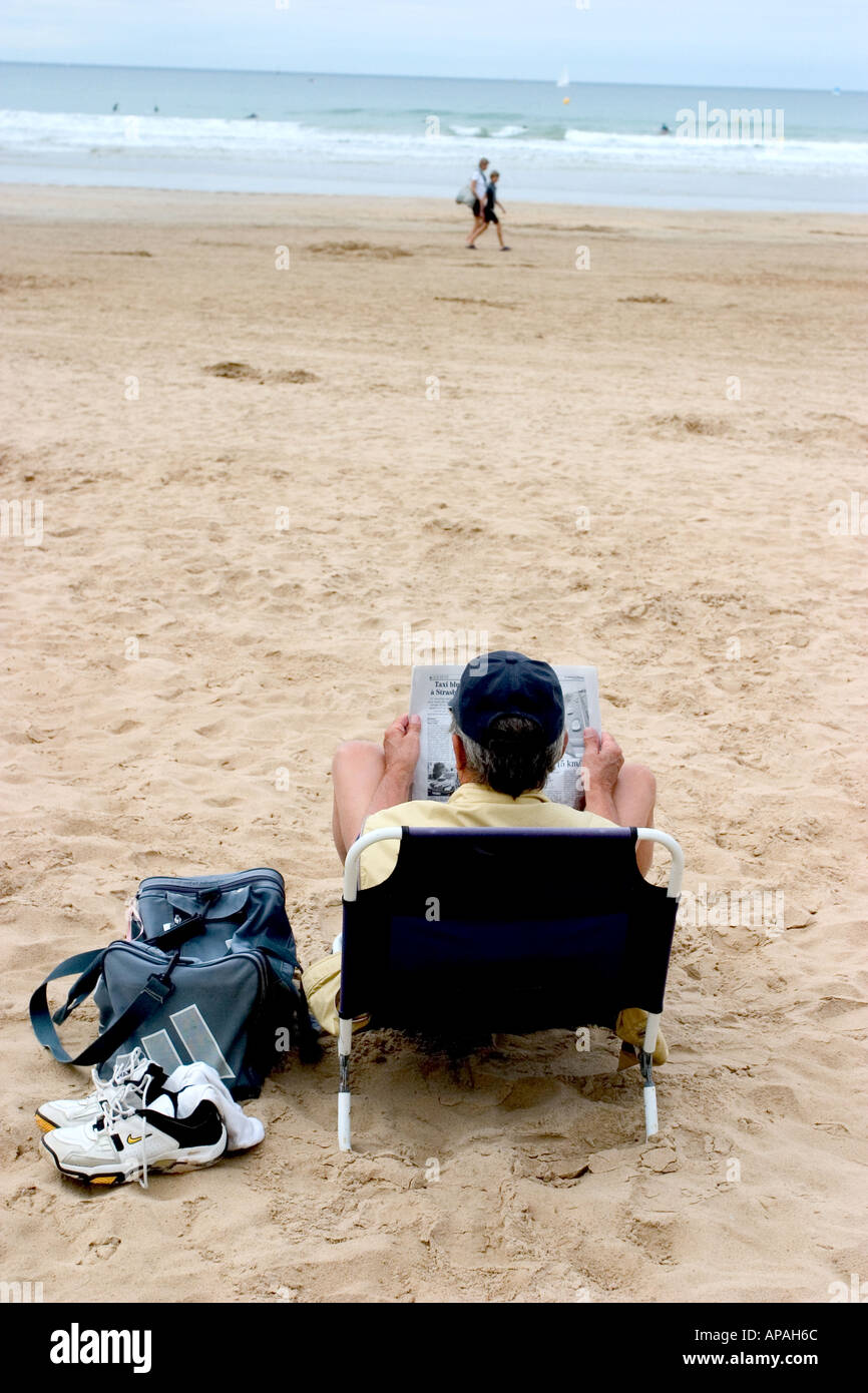 Un vieux senior man reading a magazine ou journal sur une chaise de plage en face de l'océan de la côte de la mer en france Banque D'Images