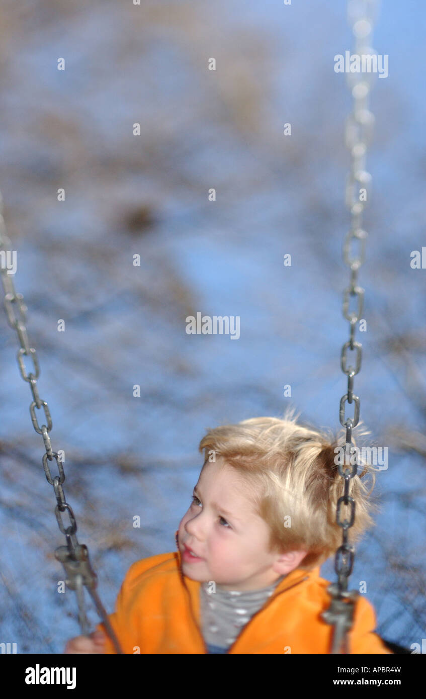 Young boy age 5-6 balançoires sur swing avec ciel bleu et les arbres et l'accent peu profondes chandail orange Banque D'Images
