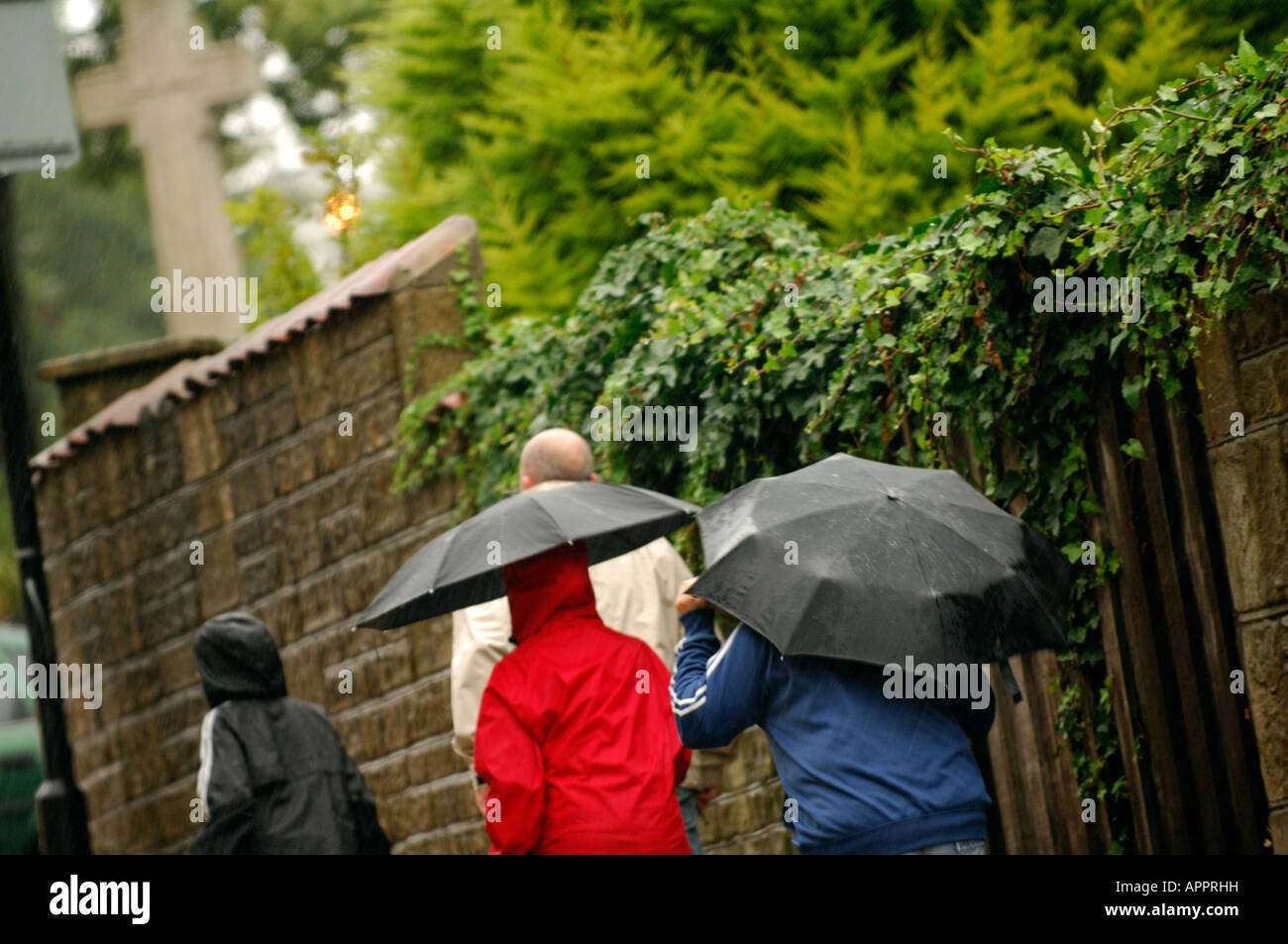 Deux personnes un couple en train de marcher le long de près d'un mur sous la pluie portant des imperméables et des parasols rouge et bleu Banque D'Images