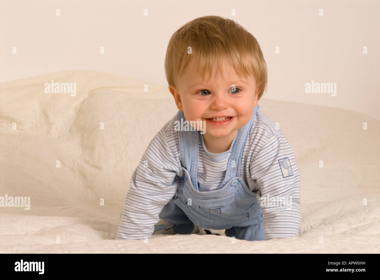 Un bébé garçon d'un an regardant la caméra rire souriant bouche heureuse ouverte habillée de T-shirt bleu et salopette sur le lit Banque D'Images