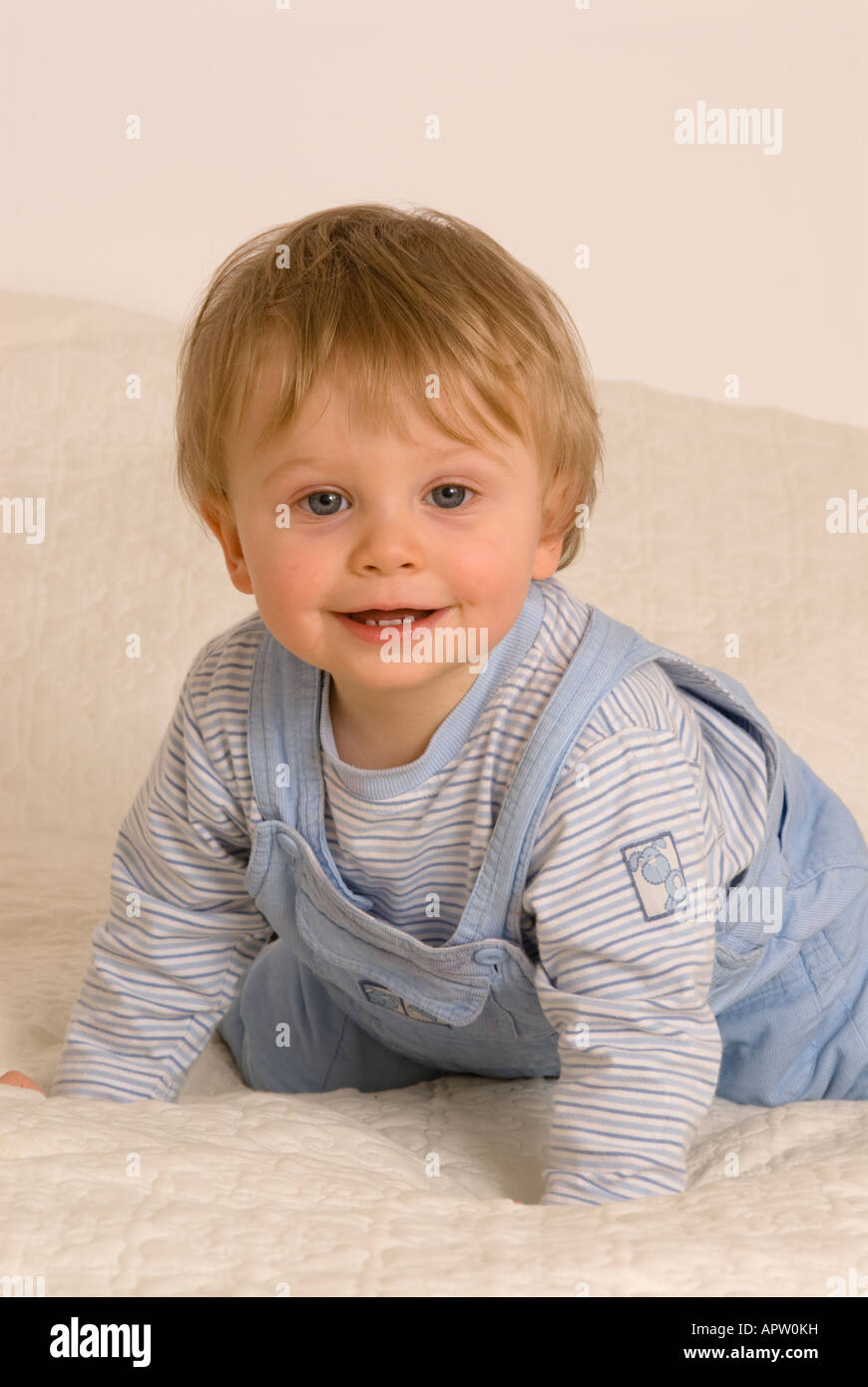 Un bébé garçon d'un an regardant la caméra rire souriant bouche heureuse ouverte habillée de T-shirt bleu et salopette sur le lit Banque D'Images