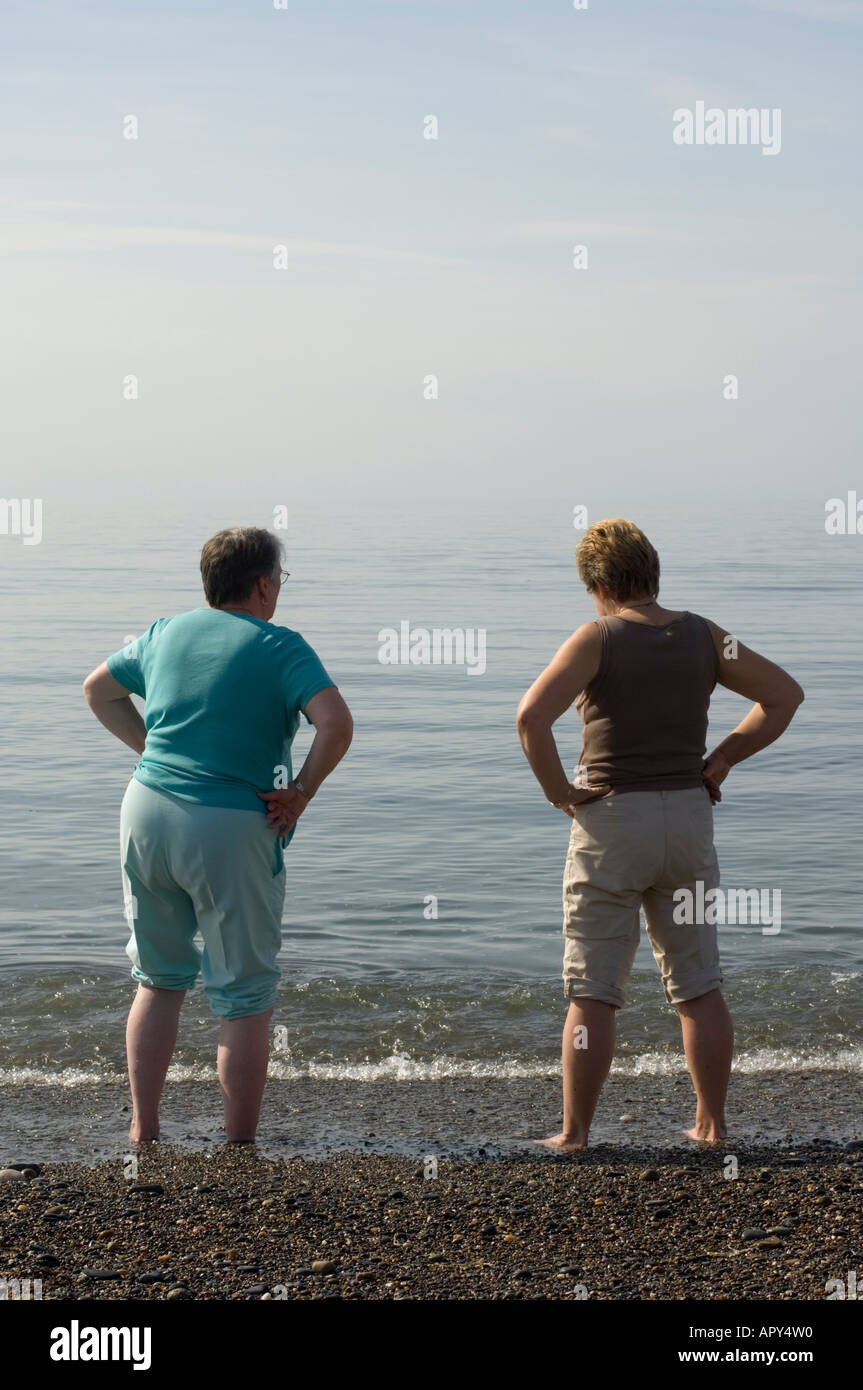 Une vue arrière de deux femmes se tenant debout avec les mains sur les hanches pagayer dans le mer plage de galles Ceredigion Aberystwyth UK après-midi d'été Banque D'Images