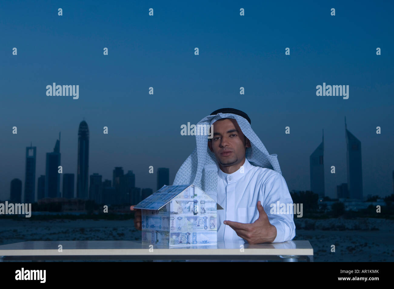 Les jeunes Arabes l'homme assis à un bureau avec la ville de Dubaï en arrière-plan la tenue d'une maison faite de l'argent Banque D'Images