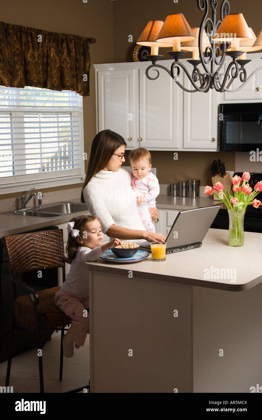 Caucasian mother holding baby et de la saisie sur ordinateur portable avec girl eating breakfast in kitchen Banque D'Images