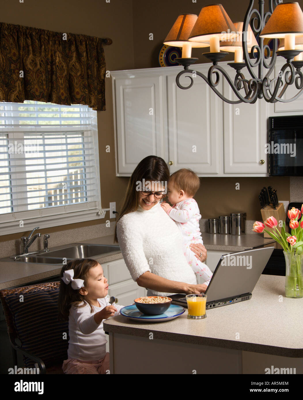 Caucasian mother holding baby et de la saisie sur ordinateur portable avec girl eating breakfast in kitchen Banque D'Images