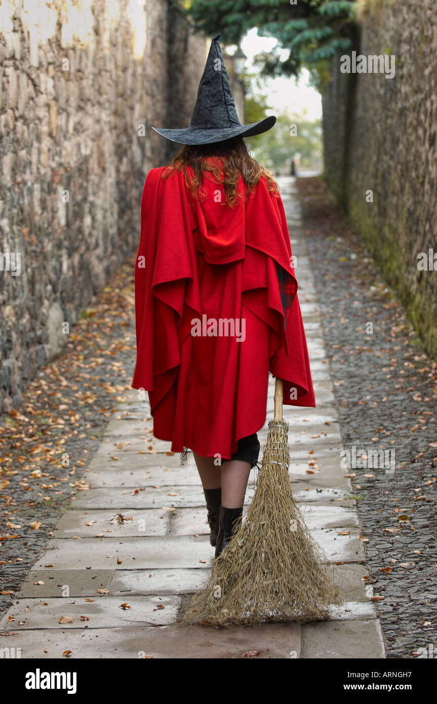 Vue arrière d'une jeune femme portant une cape rouge et un chapeau de sorcière en faisant glisser un balai marche dans une allée solitaire Banque D'Images