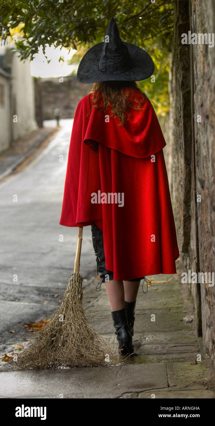 Vue arrière d'une jeune femme portant une cape rouge et un chapeau de sorcière en faisant glisser un balai marche dans une allée solitaire Banque D'Images