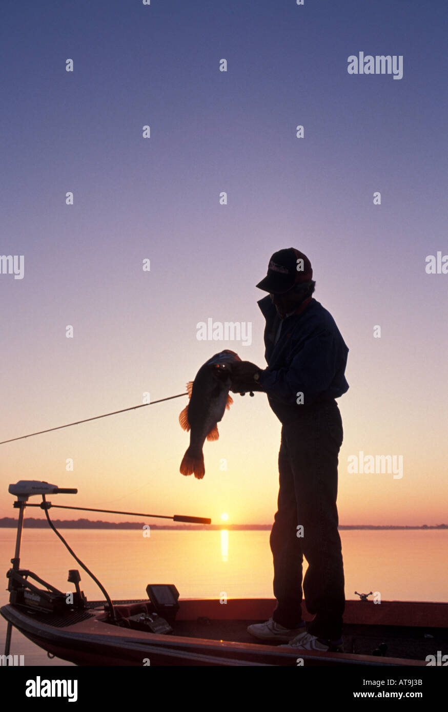 Achigan à grande bouche pêcheur pêche au lever du soleil tenant un poisson à la proue du bateau basse Banque D'Images