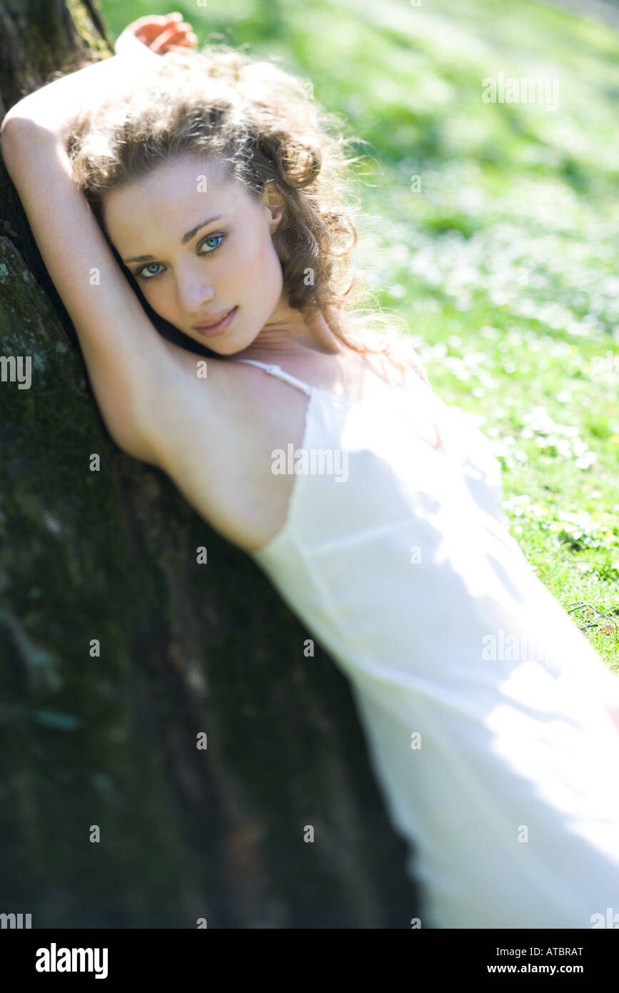 Jeune femme portant, leaning against tree trunk, bras levé, smiling at camera Banque D'Images