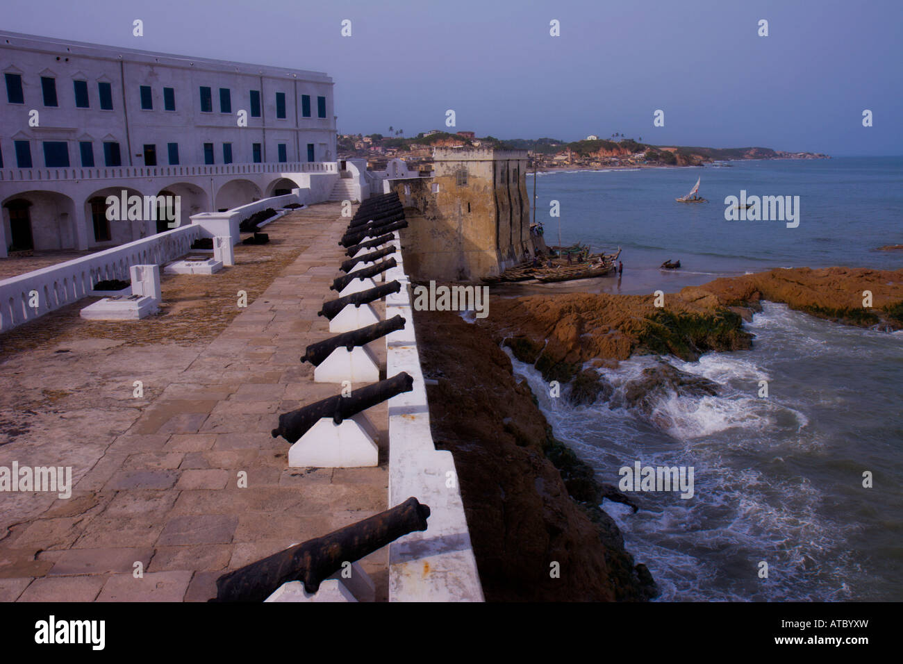 Une série de canons sur les murs du château de Cape Coast signaler par la mer, tandis que les vagues déferlent sur les rochers et d'un bateau navigue dans le port. Banque D'Images