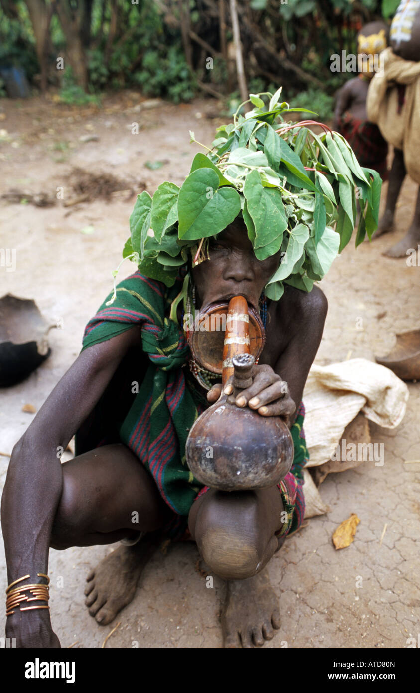 Une vieille femme Surma bénéficie d'une fumée traditionnelle à côté de sa cabane dans son village Banque D'Images