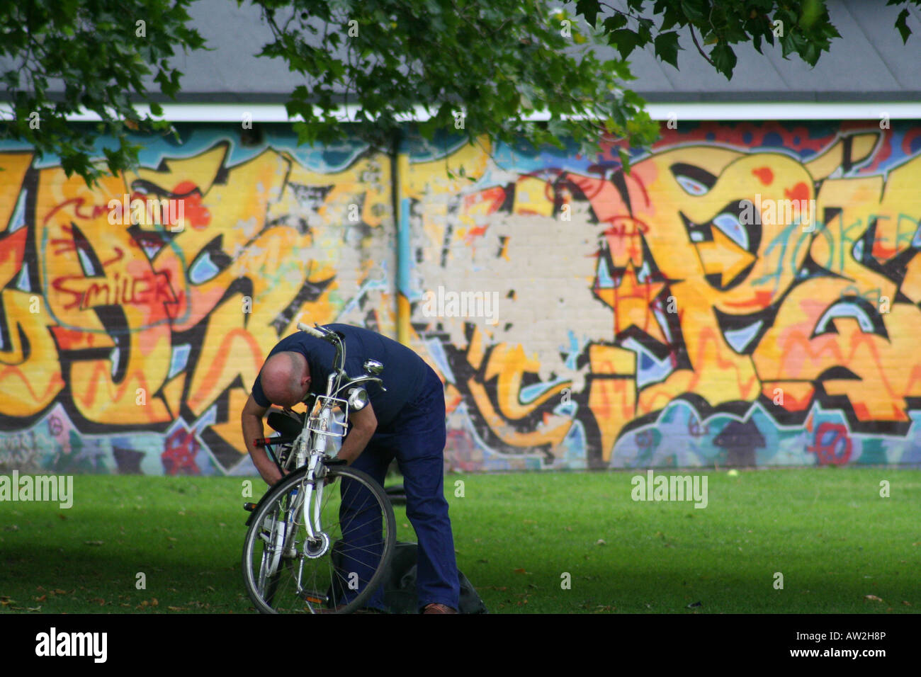 Une tête chauve homme habillé en bleu ajuste son vélo dans un parc de Londres devant un mur couvert de graffitis Banque D'Images