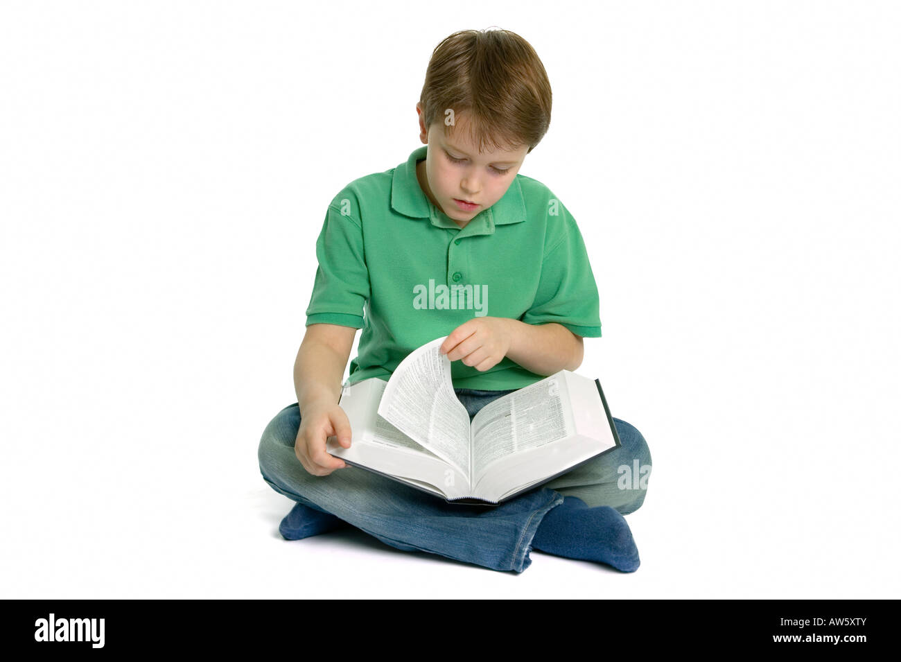 Un jeune garçon est assis jambes croisées en tournant la page d'un ouvrage de référence il lit tourné sur un fond blanc. Banque D'Images