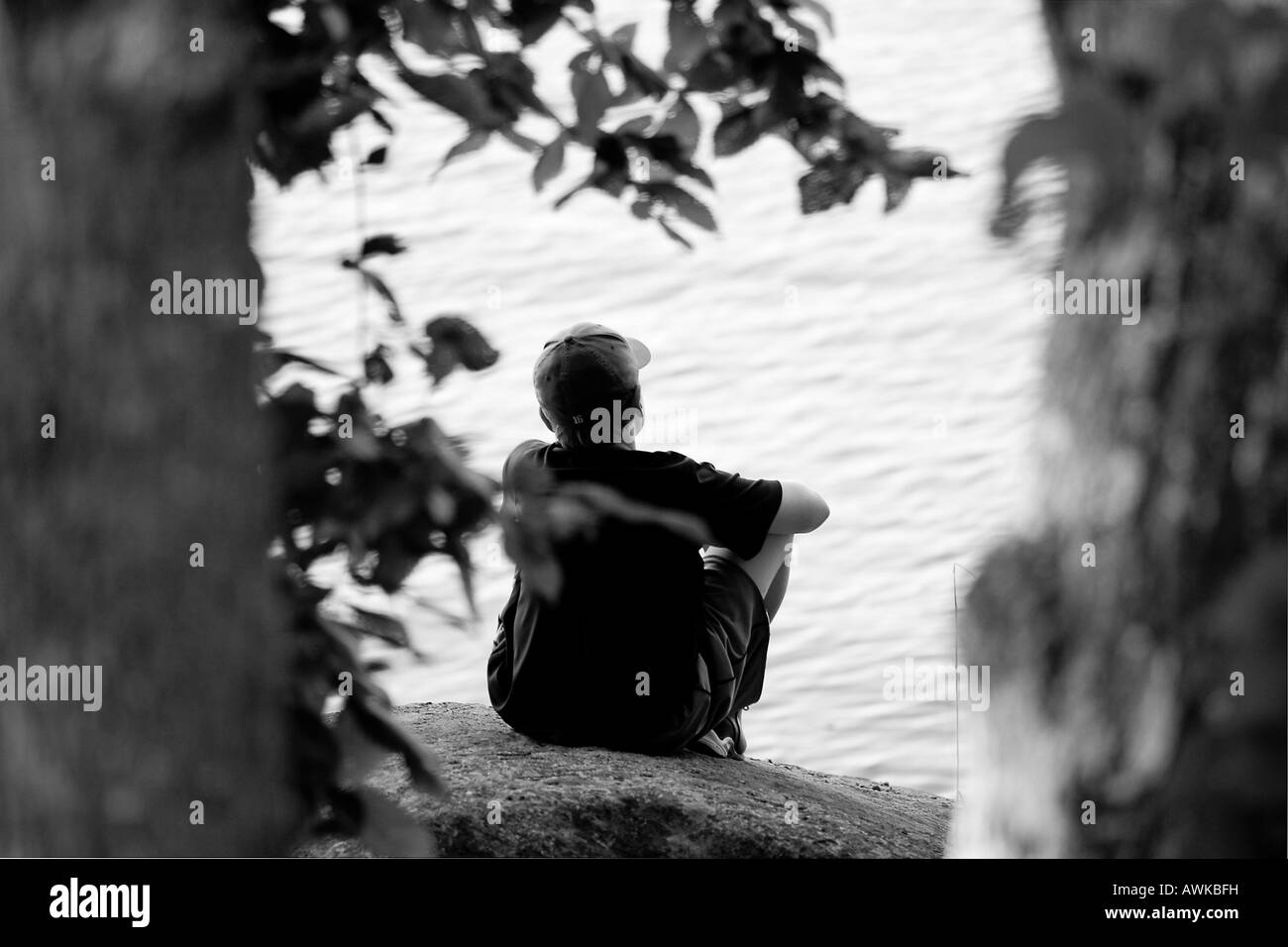 Une image en noir et blanc d'un jeune garçon est assis sur un rocher sur le rivage d'un lac, entouré par de grands arbres. Banque D'Images