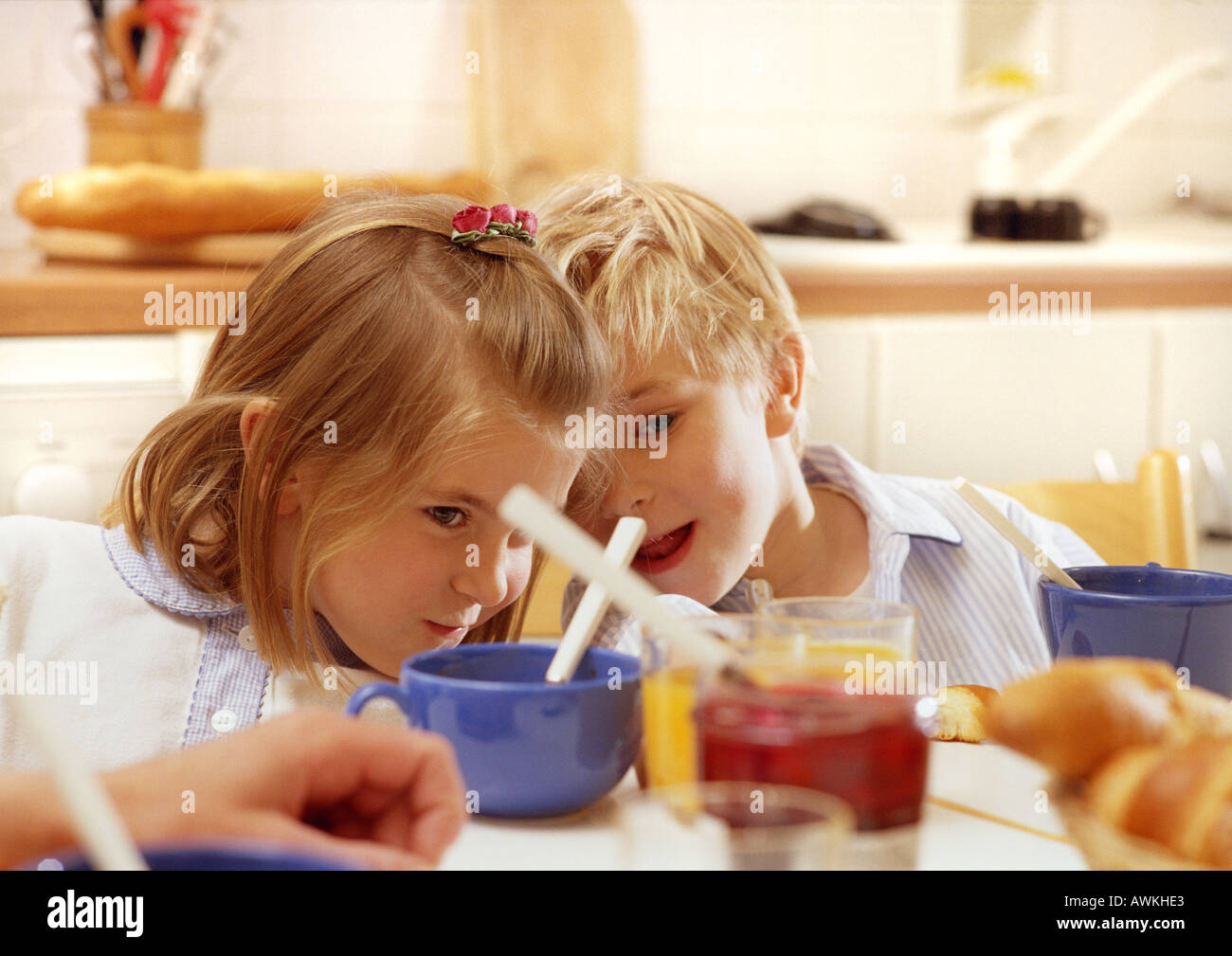 Des enfants assis à table leaning chefs ensemble, tête et épaules, tasses, pot de gelée et floue main adultes en premier plan Banque D'Images