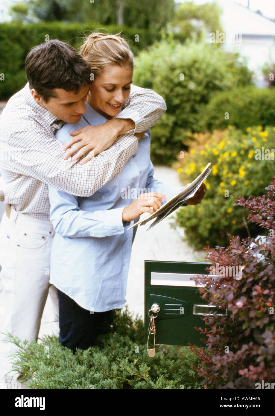 Woman holding mail, l'homme l'étreindre par derrière, à côté de la boîte de courrier électronique, des arbustes en arrière-plan Banque D'Images