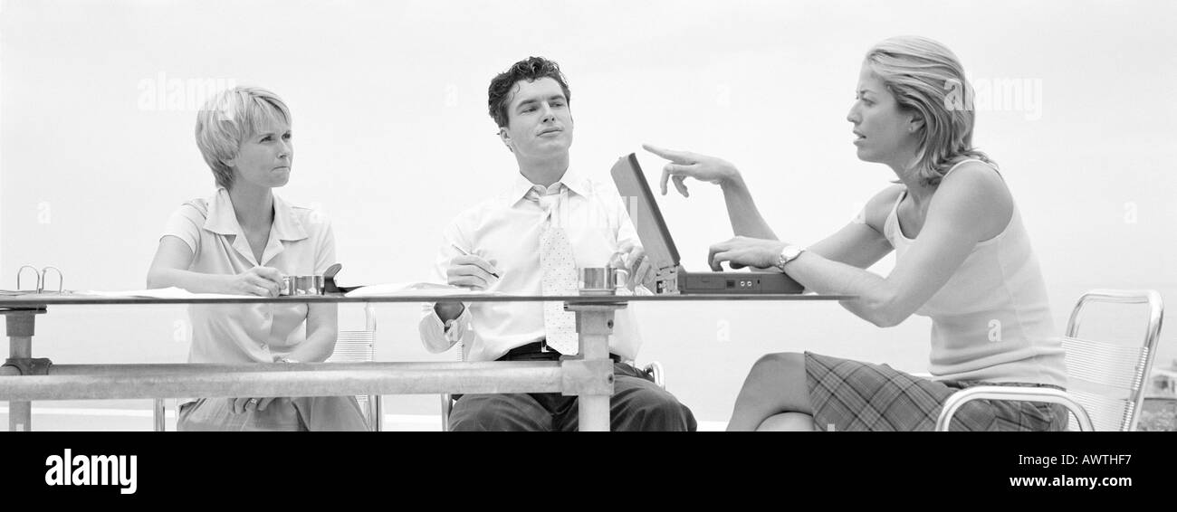 Trois personnes assises à table, l'un parlant et en pointant sur l'écran du portable, b&w, vue panoramique Banque D'Images