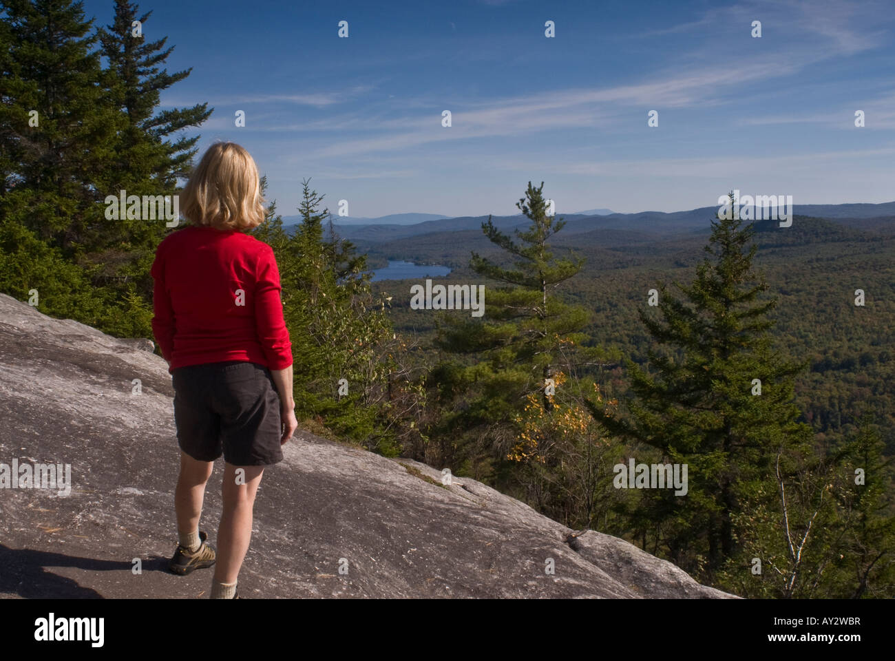 Une femme dans un tee shirt rouge debout sur une saillie rocheuse au sommet le Mont Owl's Head en regardant le lac et les montagnes au loin. Banque D'Images