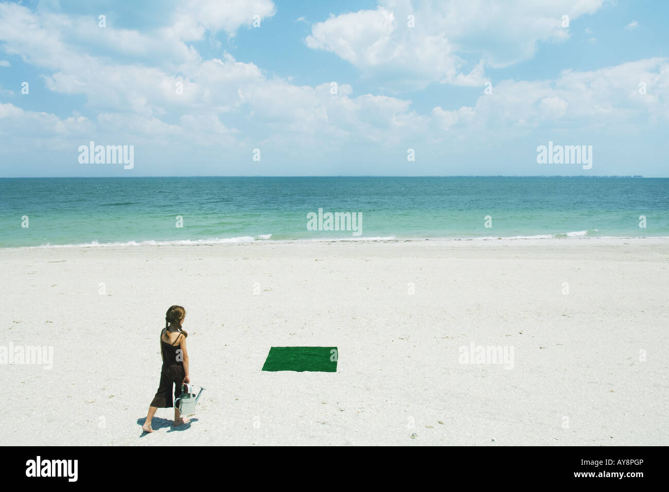 Fille à la plage à pied en direction de patch de gazon artificiel, exerçant son arrosoir, vue arrière Banque D'Images