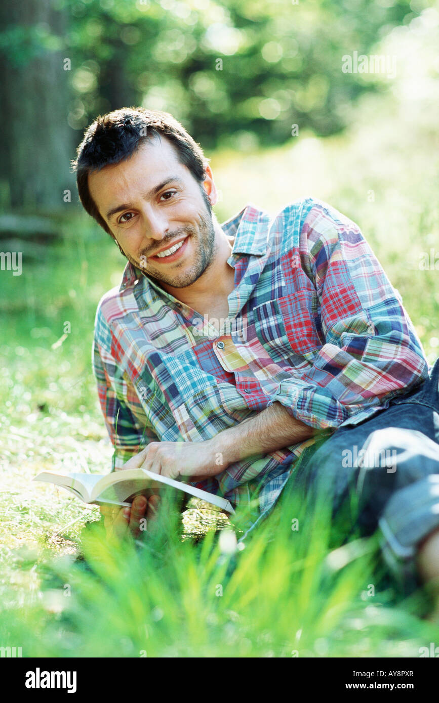 Homme assis sur le sol à l'extérieur, holding book, smiling at camera Banque D'Images