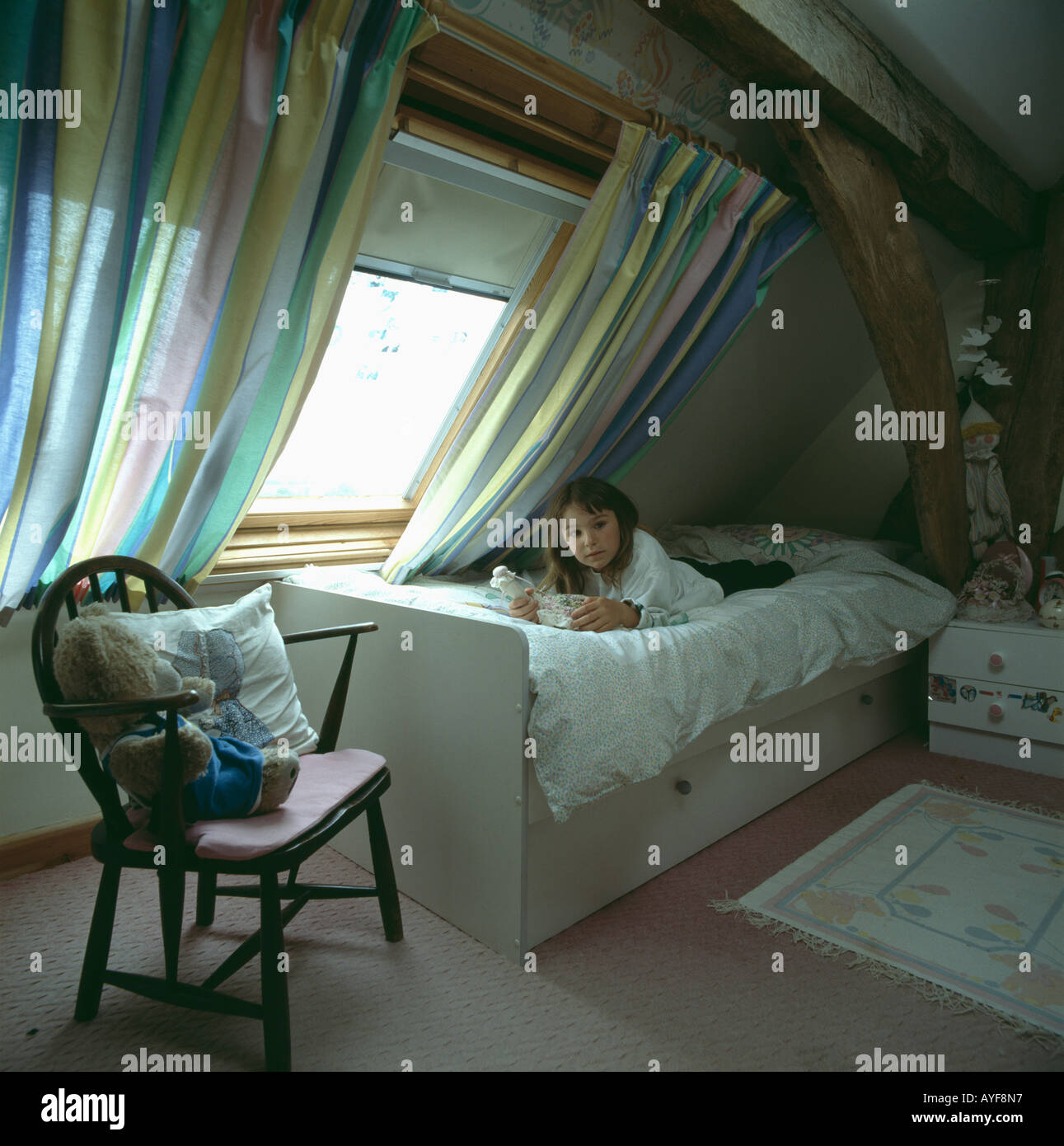 Petite girl lying on bed et de la lecture ci-dessous fenêtre avec rideaux rayés dans une chambre mansardée Banque D'Images