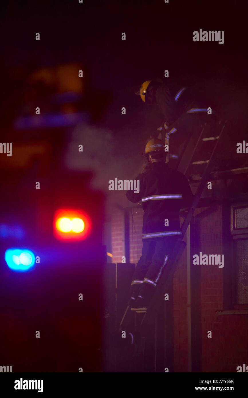 pompiers au sommet d'une échelle sur le toit d'un bungalow combattant un feu de maison domestique dans un Rue résidentielle au Royaume-Uni en supprimant les tuiles de toit Banque D'Images