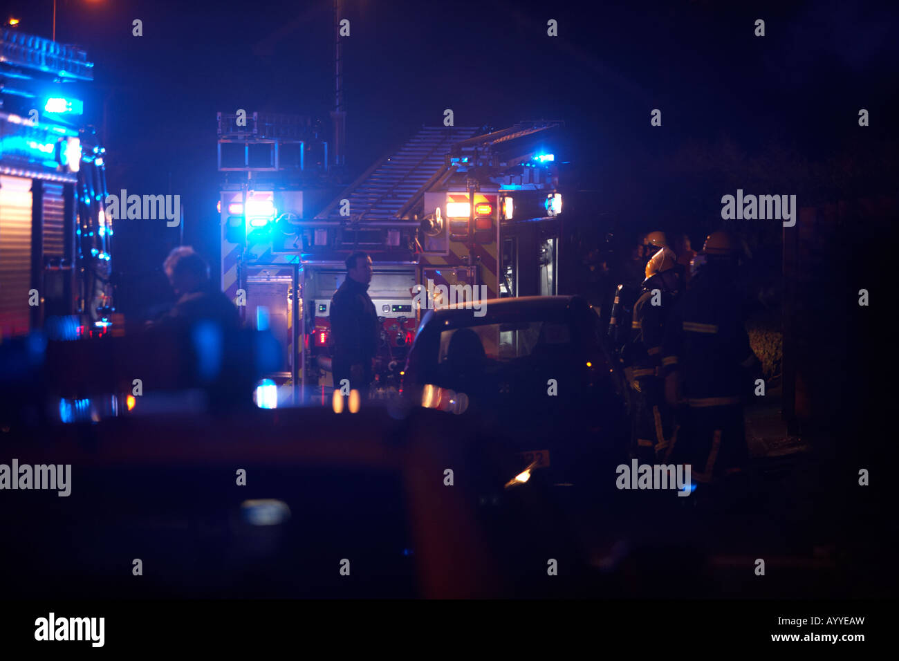 Les pompiers en attente derrière un camion d'incendie moteur de pompage par des voitures en stationnement sur les lieux de l'incendie d'une maison au Royaume-Uni Banque D'Images