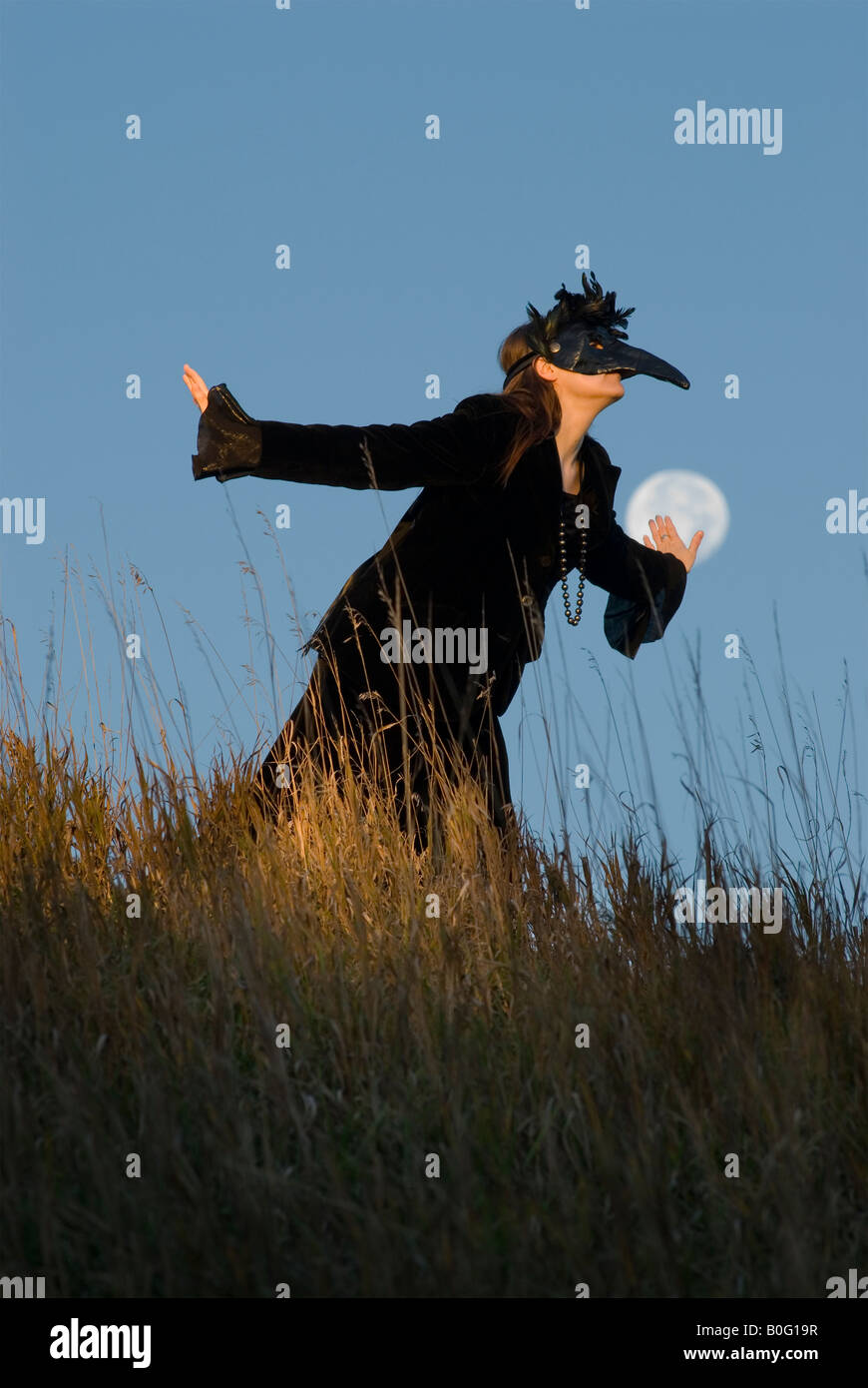 Une femme masquée dans une robe de velours noir se présente comme un corbeau ou Raven Spirit avec la pleine lune en arrière-plan Banque D'Images