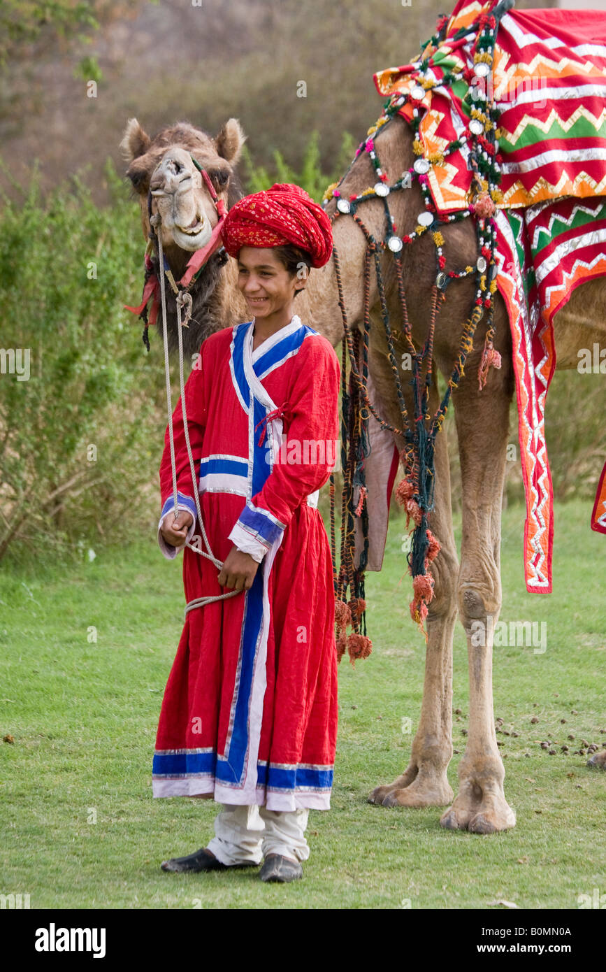 East Indian boy stands smiling vêtus de couleur rouge brillant costume de cérémonie ethnique tenant un chameau décorées de couleurs vives Jaipur Rajasthan Inde Banque D'Images