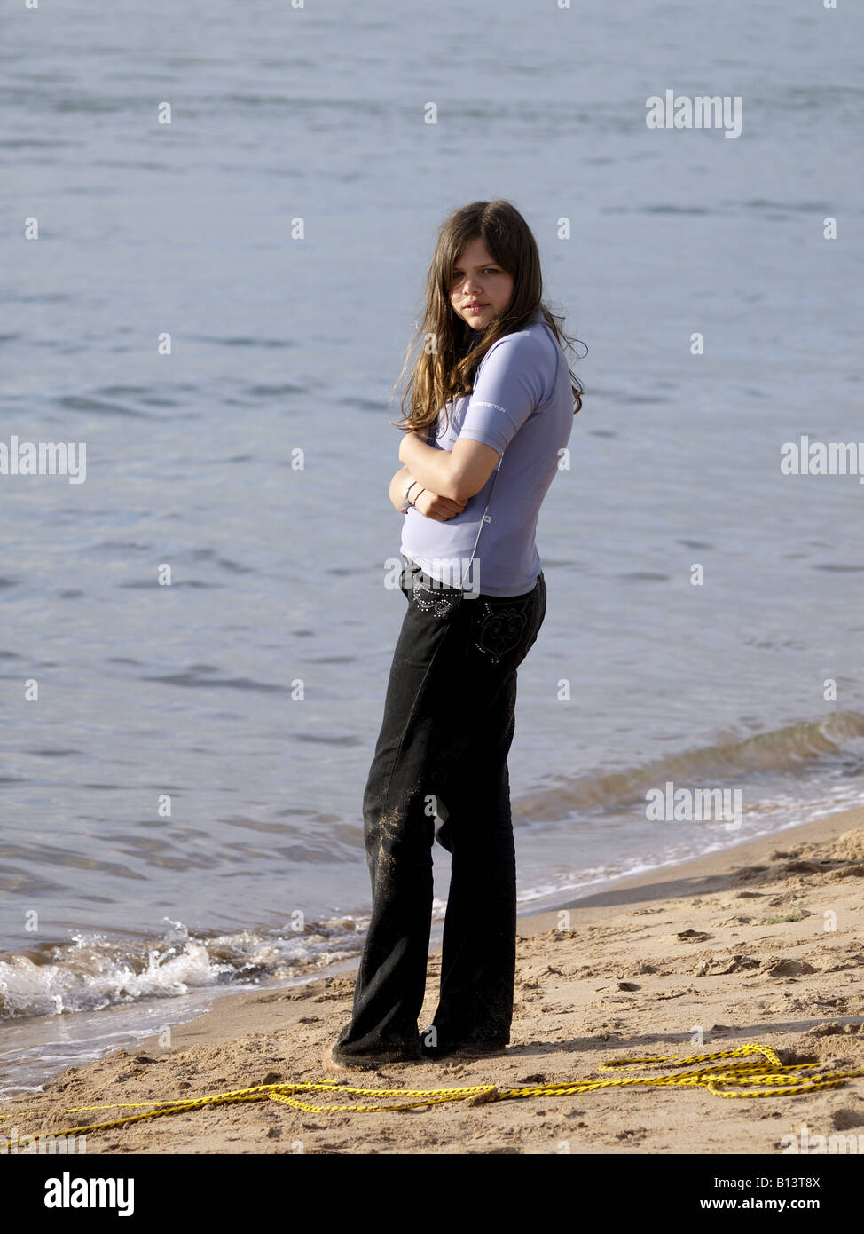 Adolescente avec tout droit, long, cheveux bruns, portant une paire de jeans noir, debout à côté de la mer avec les bras croisés Banque D'Images